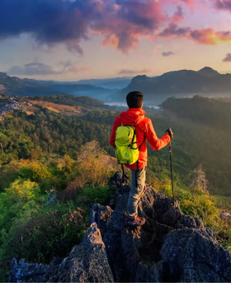 Hiker in a red jacket and yellow backpack standing on a mountain peak at sunset