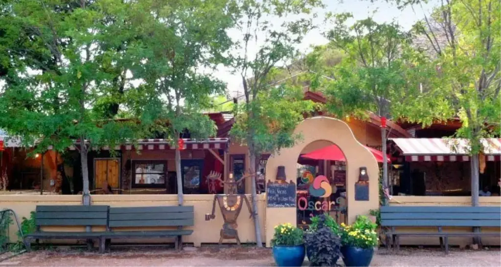 Exterior view of Oscar's restaurant featuring a southwestern-style arched entrance, outdoor patio with red umbrellas, and rustic green wooden benches under shade trees.