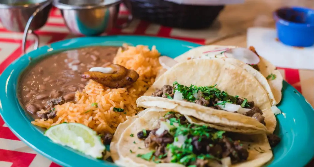 Mexican food platter with three street tacos, seasoned rice, refried beans, and a lime wedge on a bright turquoise plate